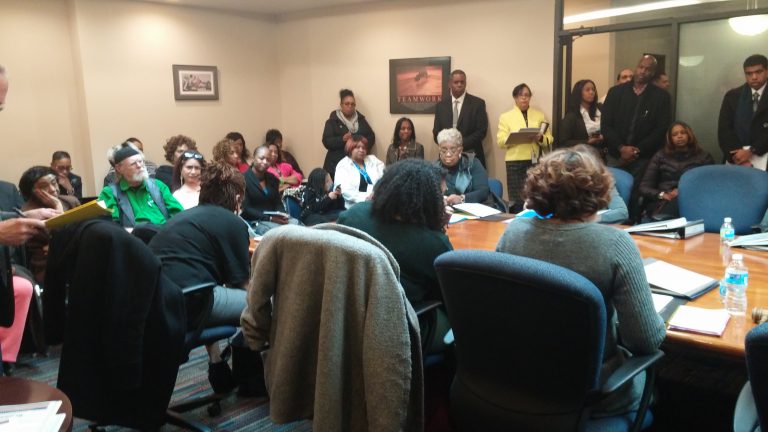 A city government meeting around a large conference table. The room is full of people standing and sitting.