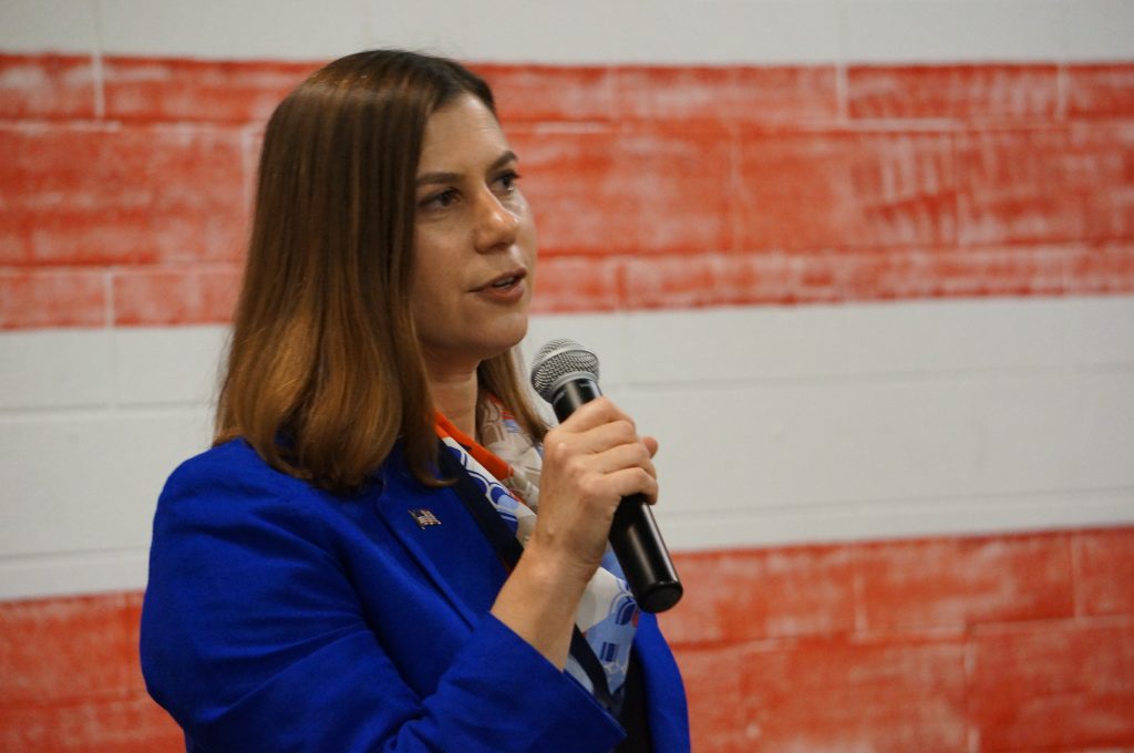 a woman in a blue blazer speaks into a microphone in front of an American flag