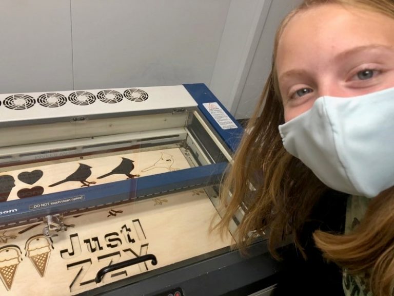 a teenage girl wearing a protective face mask smiles in front of a machine with wooden cutouts of figures like birds, hearts and ice cream cones