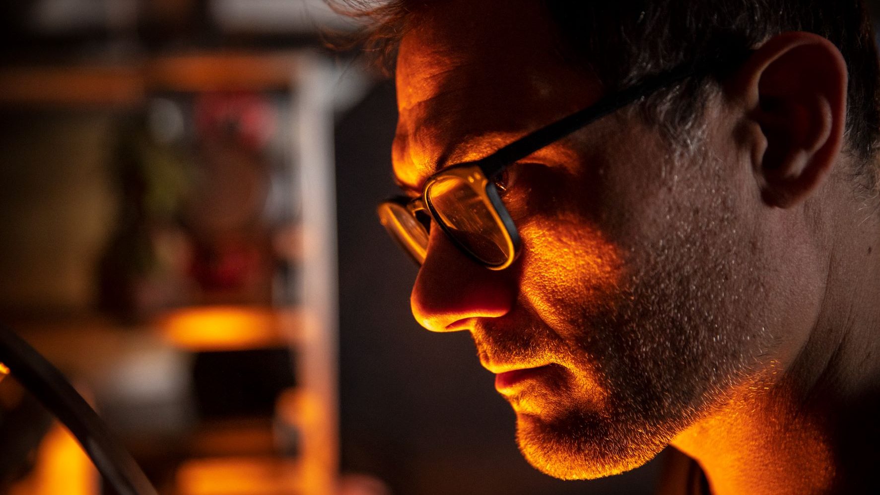 close-up of a man wearing glasses, whose face glows orange from a nearby wood-fired oven