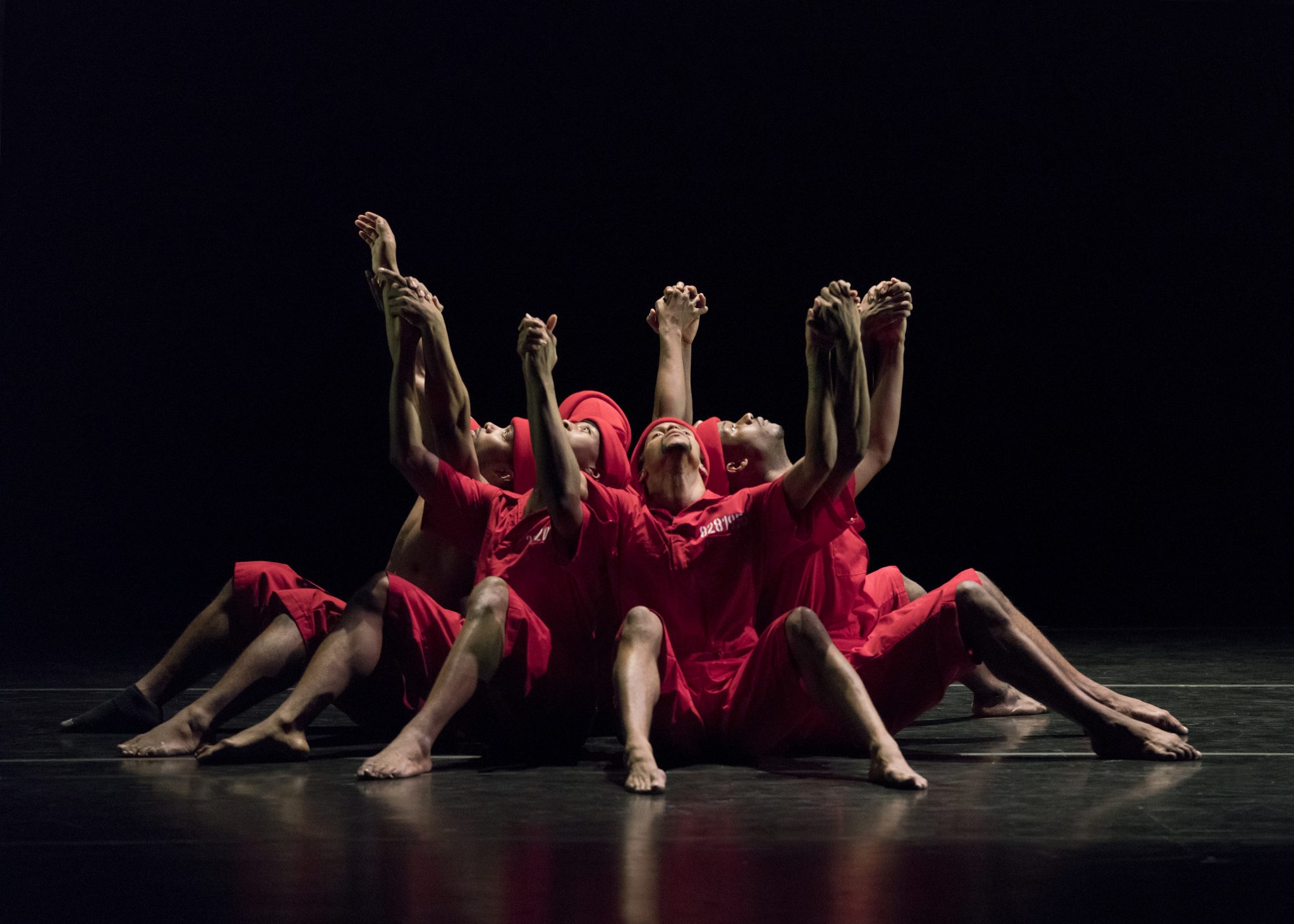 a group of black men in red costumes sit in a circle on stage with their hands above their heads