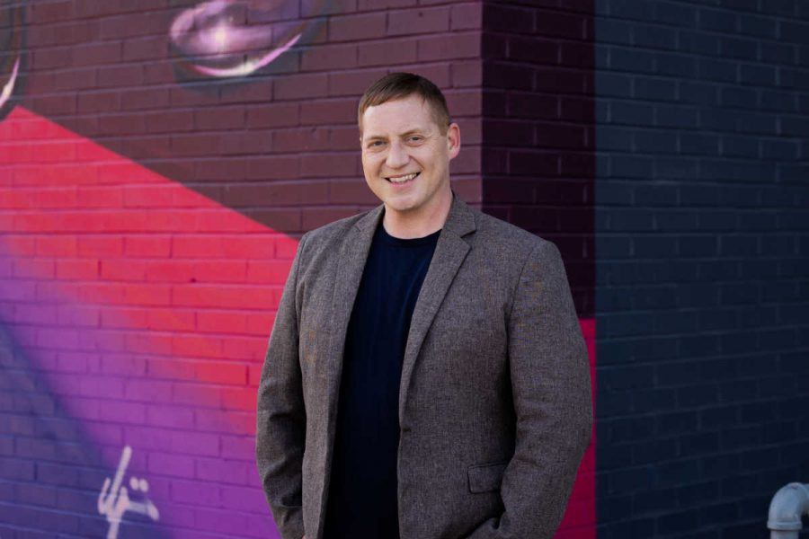 a man in a suit coat smiles in front of a pink, purple and blue mural