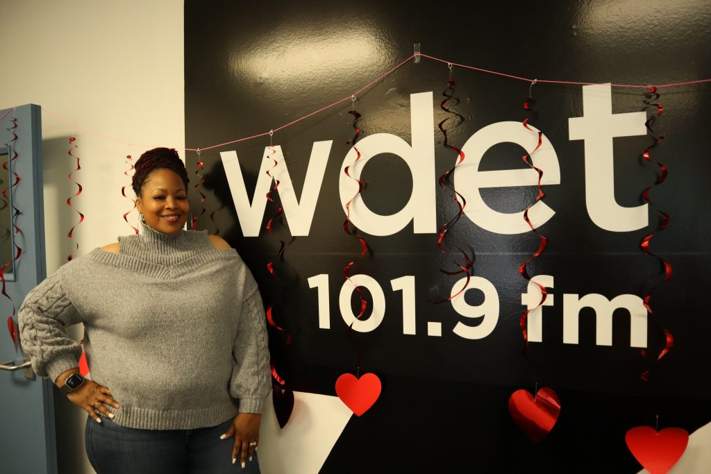 a Black woman in a stylish gray sweater poses in front of the WDET logo