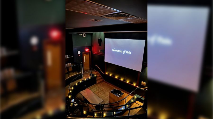 a dark auditorium lit by candles and a screen that reads "Narratives of Pain"