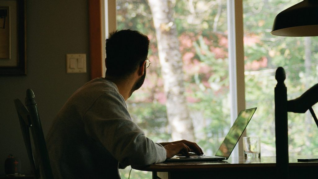 man types on a laptop and looks out a window