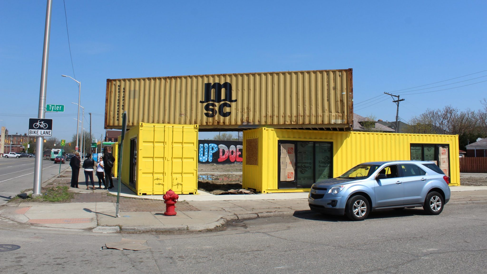 A shipping container is stacked on top of two bright yellow shipping containers. Behind them you can see part of a mural that says "What Up Doe."