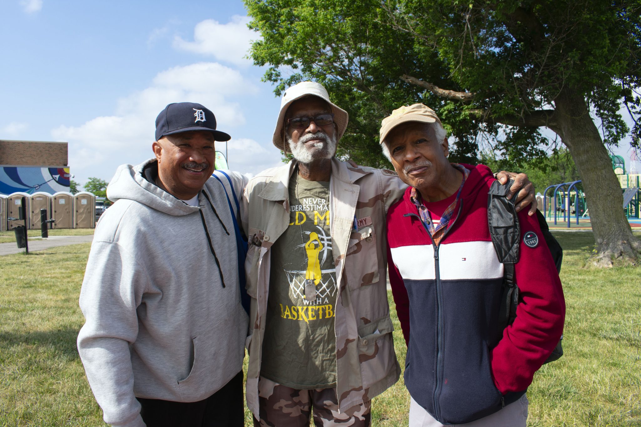 Three older Black men smile outside with their arms around each other