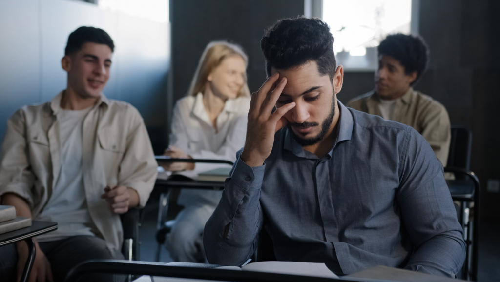 A male student sits in class at a desk alone with his head in his hand, while other students socialize behind him.