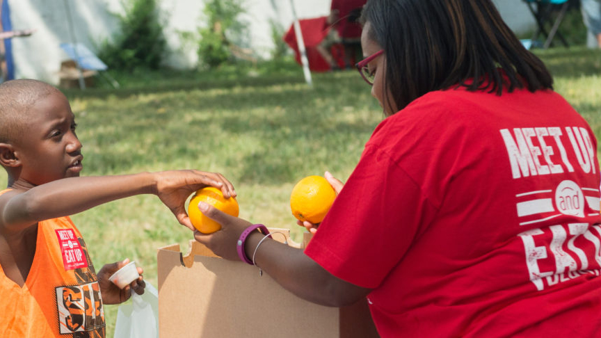 A volunteer hands a child an orange during a Meet Up and Eat Up event.