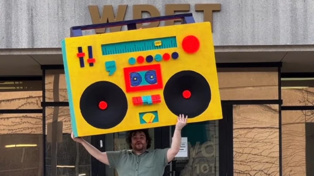 In The Groove host Ryan Patrick Hooper holds Detroit's largest boombox — designed by Detroit artist Matthew Holman — outside of WDET Studios during the 2025 spring fundraiser.