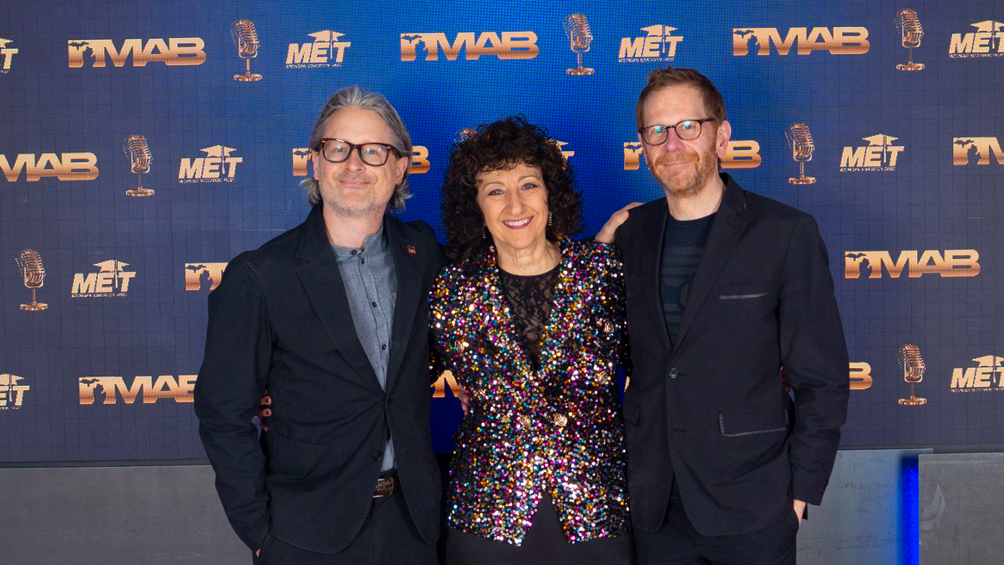WDET staff member and hosts stand in front of the MAB Step and Repeat