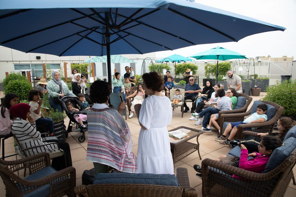 Children and adults sit in a large circle in a rooftop garden with patio furniture, raised bed gardens, and umbrellas for shade.