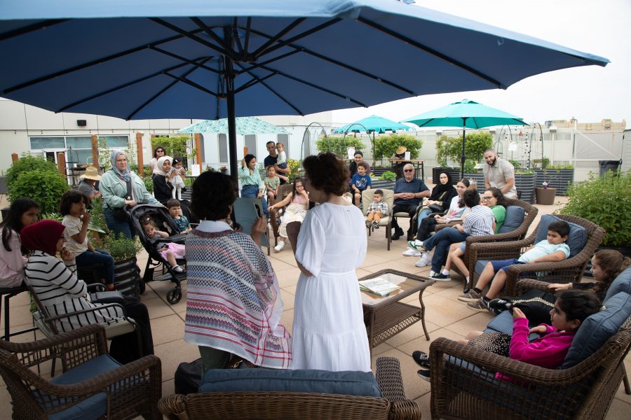 Children and adults sit in a large circle in a rooftop garden with patio furniture, raised bed gardens, and umbrellas for shade.