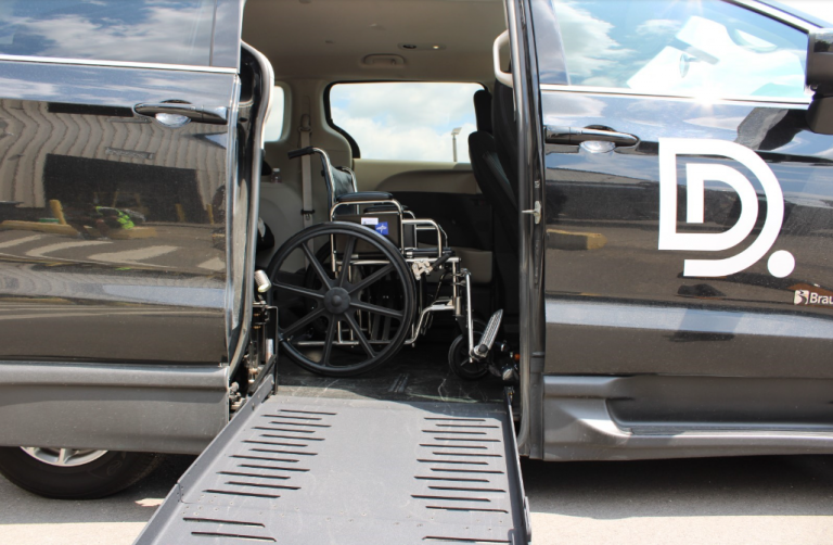 A mini van is parked with its side door open. A ramp is coming out the side and you can see a wheelchair in the van.