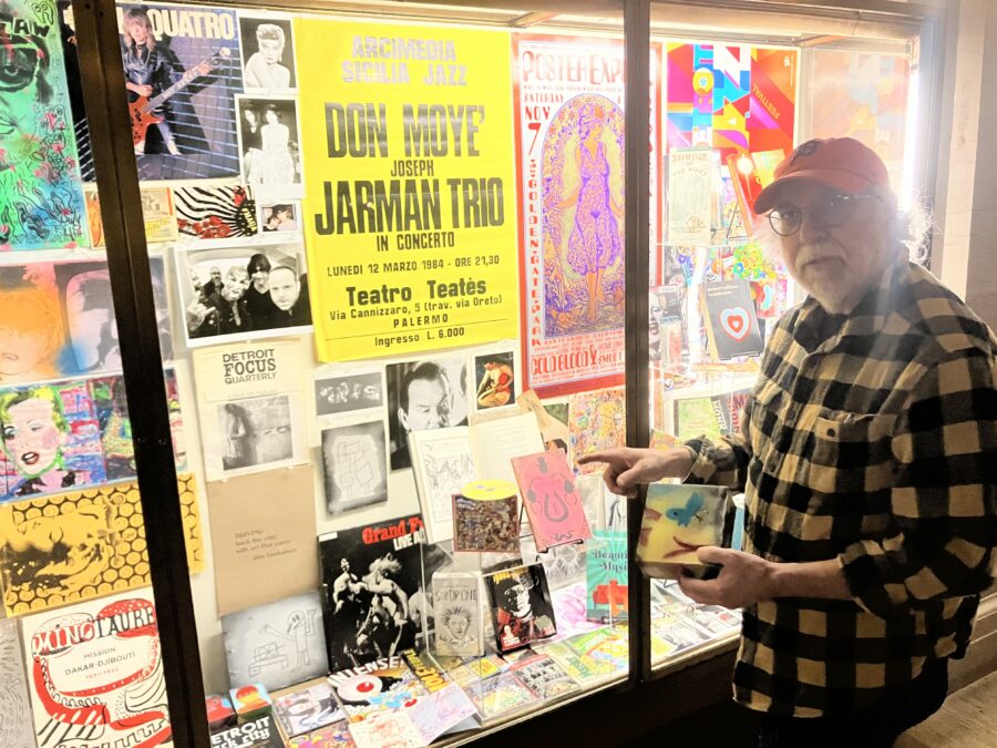 A man wearing flannel and a baseball cap stands in front of a colorful display of magazines, posters, and visual ephemera.