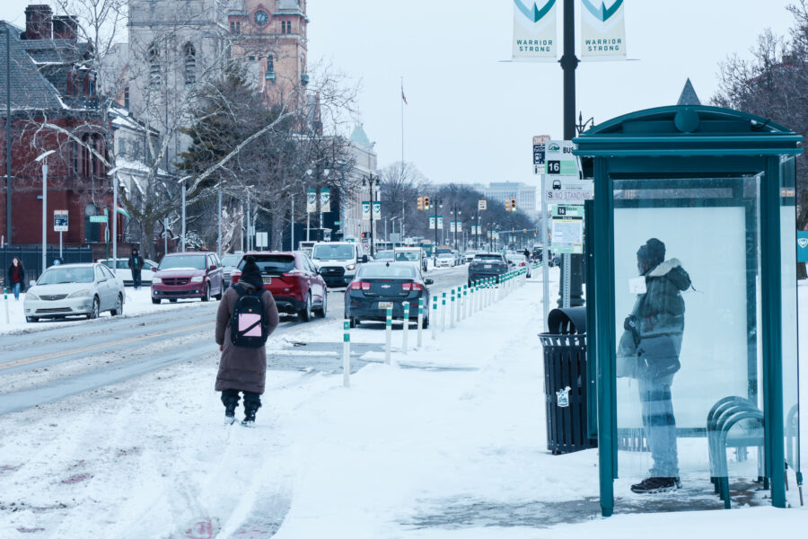 A person in winter gear waits to safely pass a snow-covered street in Midtown Detroit. Another individual stands in the bus stop shelter.