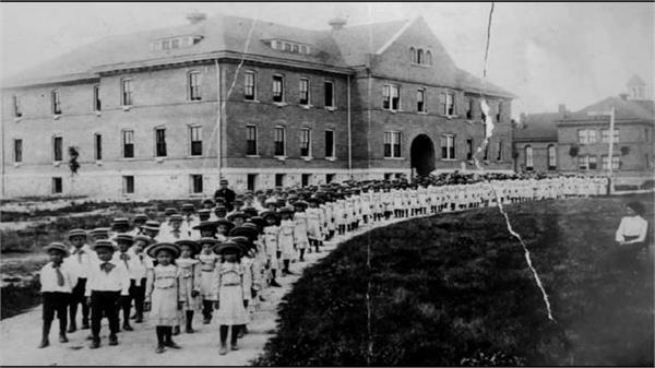 Dozens of "Americanized" native children stand posed in front of a boarding school, dressed in the dominant fashion of the time.