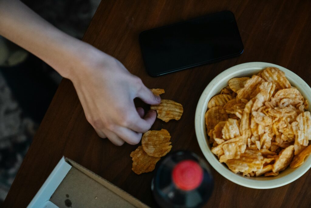Hand reaching for potato chips strewn on a table next to a bottle of soda.