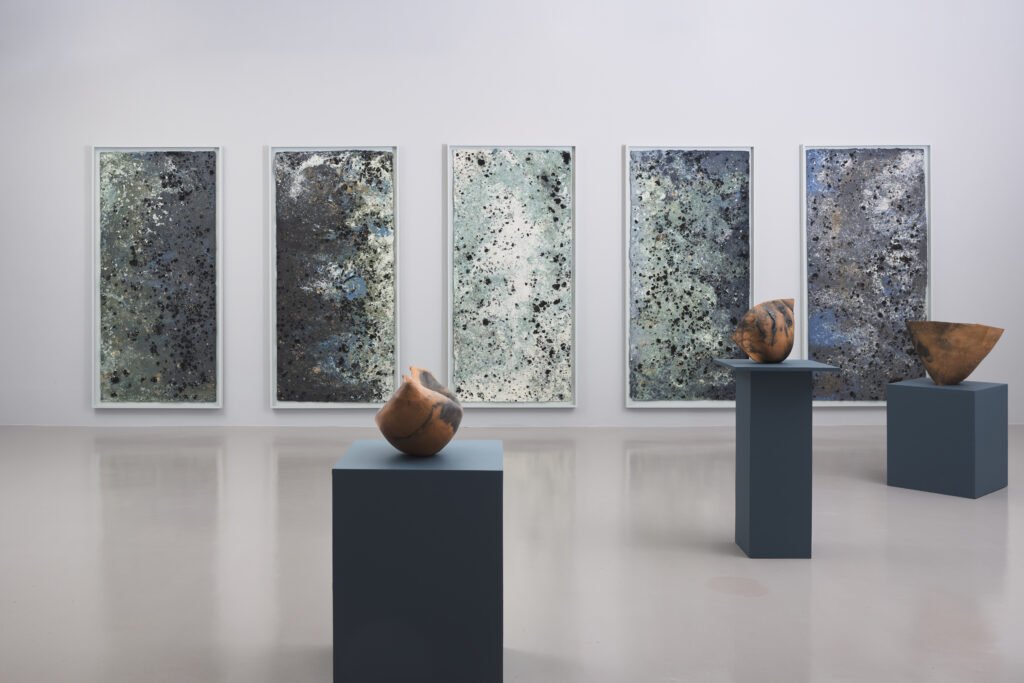 An art gallery showing tall canvasses of blue paint. Vases rest on columns in front of the paintings.