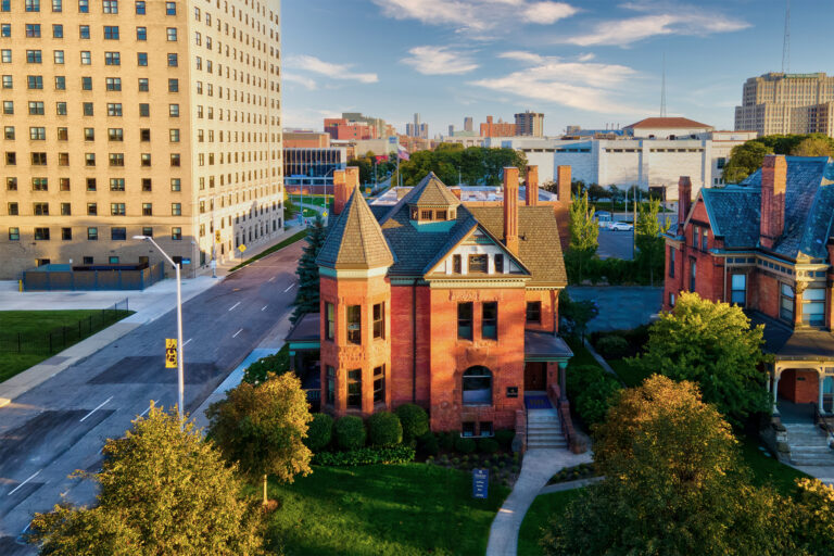 A red brick house stands in Midtown, with a skyscraper across the street and trees in the foreground.