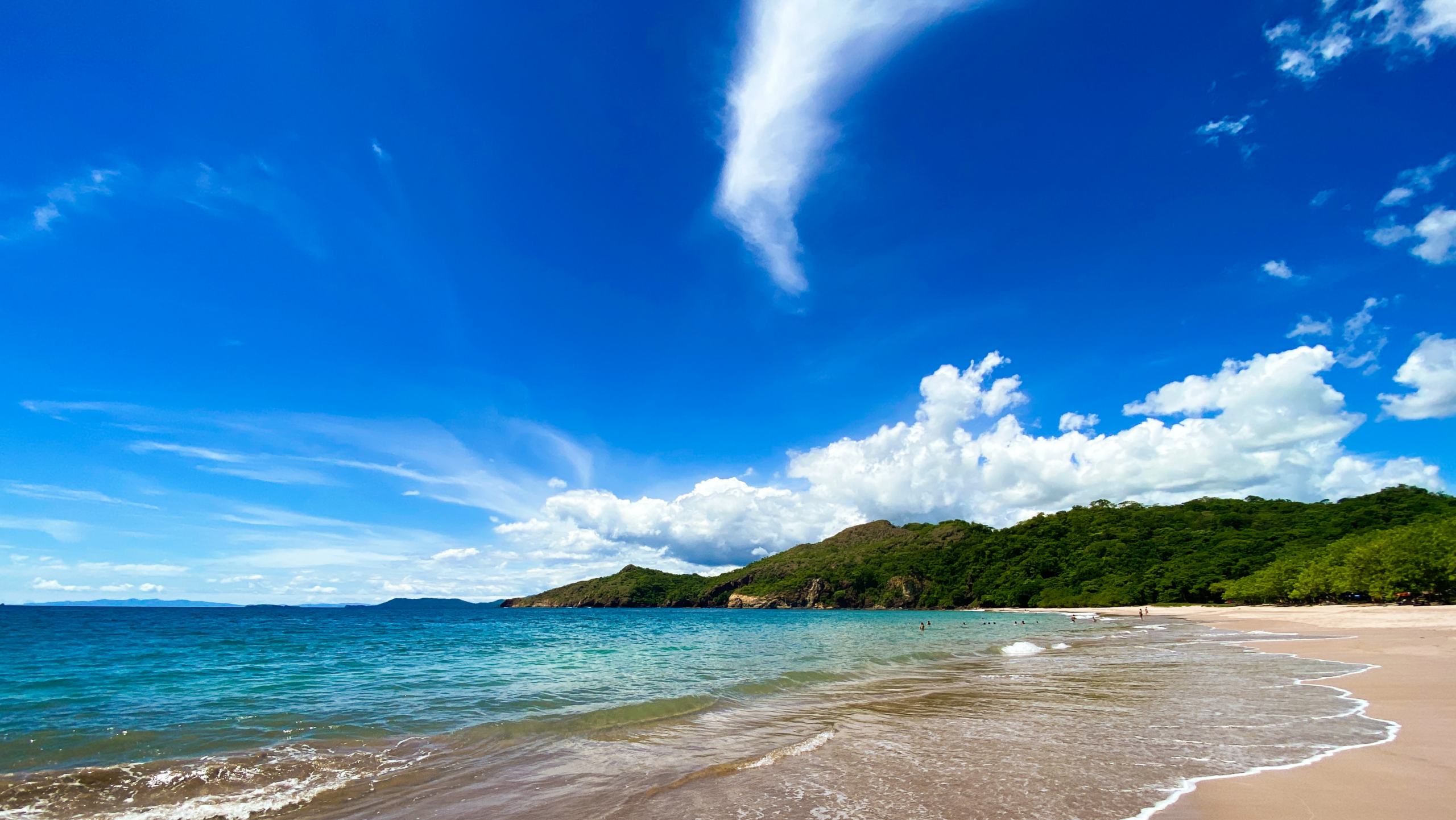 Sunny tropical beach with gentle waves washing onto a sandy shoreline, clear blue water, and lush green hills under a bright blue sky with scattered clouds.
