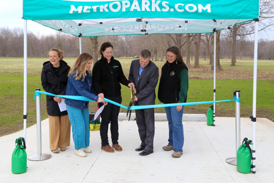 A man with large shears and 4 other people are standing at a campground and cutting the ribbon on a pop up tent labeled metroparks.com.