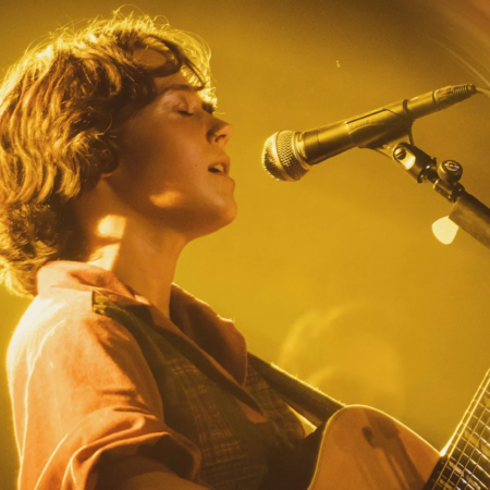 A women is singing into a microphone while playing guitar. She is on stage with hazy lighting.