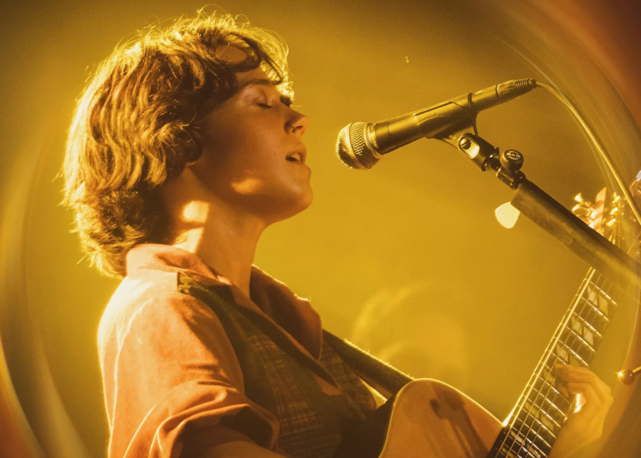 A women is singing into a microphone while playing guitar. She is on stage with hazy lighting.