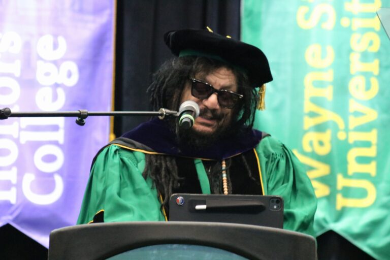 Don Was in green Wayne State doctoral robes speaks into the microphone on stage.