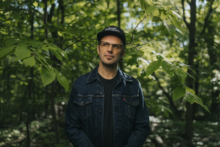 Mark Elleri stands in the woods looking pensively off to the side. He wears a baseball cap, glasses and a jean jacket over a black t-shirt.