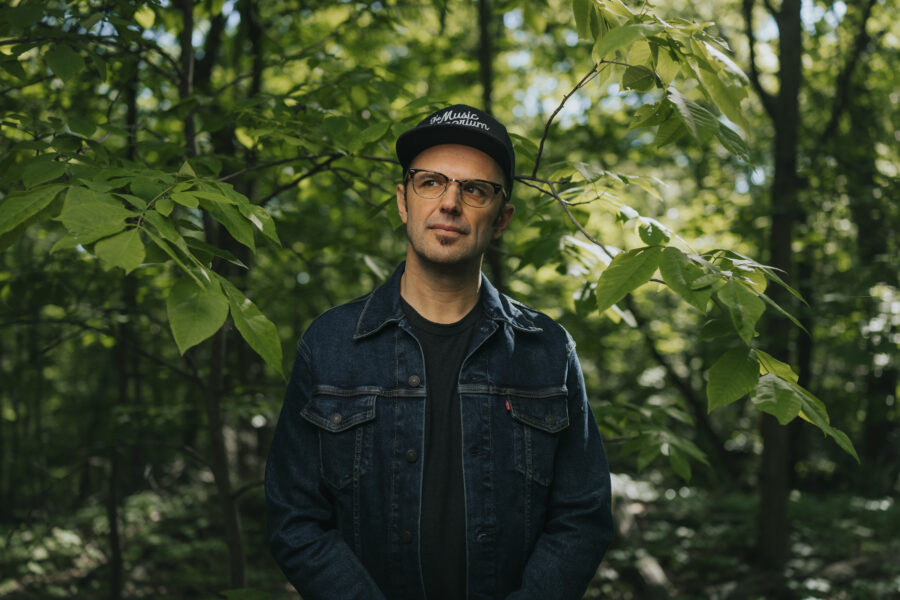 Mark Elleri stands in the woods looking pensively off to the side. He wears a baseball cap, glasses and a jean jacket over a black t-shirt.