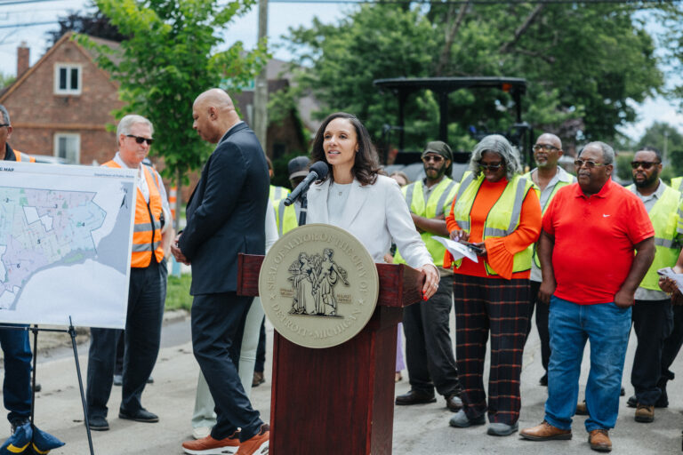 Mayor Sheffield stands at a podium in front of people wearing high-visibility vests commonly worn by construction workers.