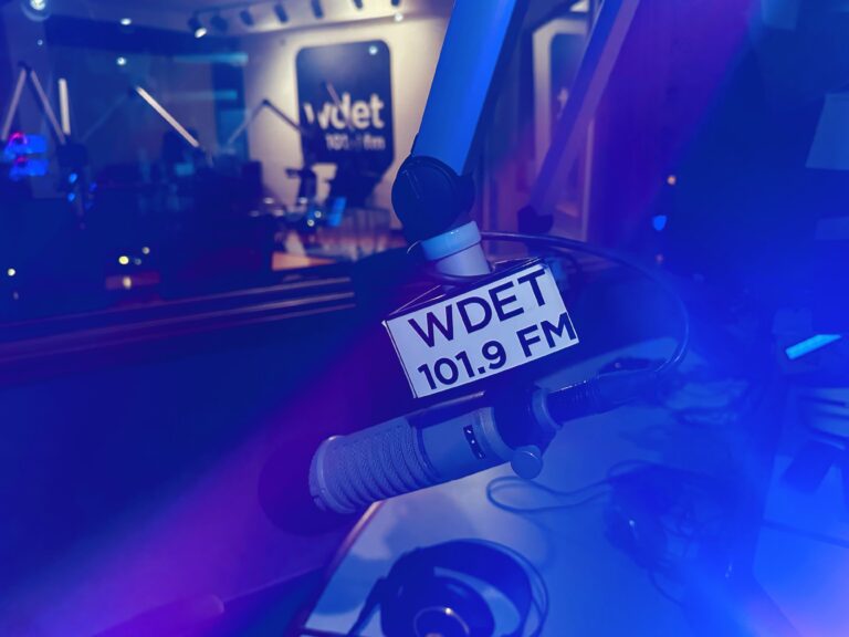 A purple-lit WDET studio mic. A white flag on the mic reads "WDET 101.9 FM" in black text. In the background, a WDET logo is lit up.