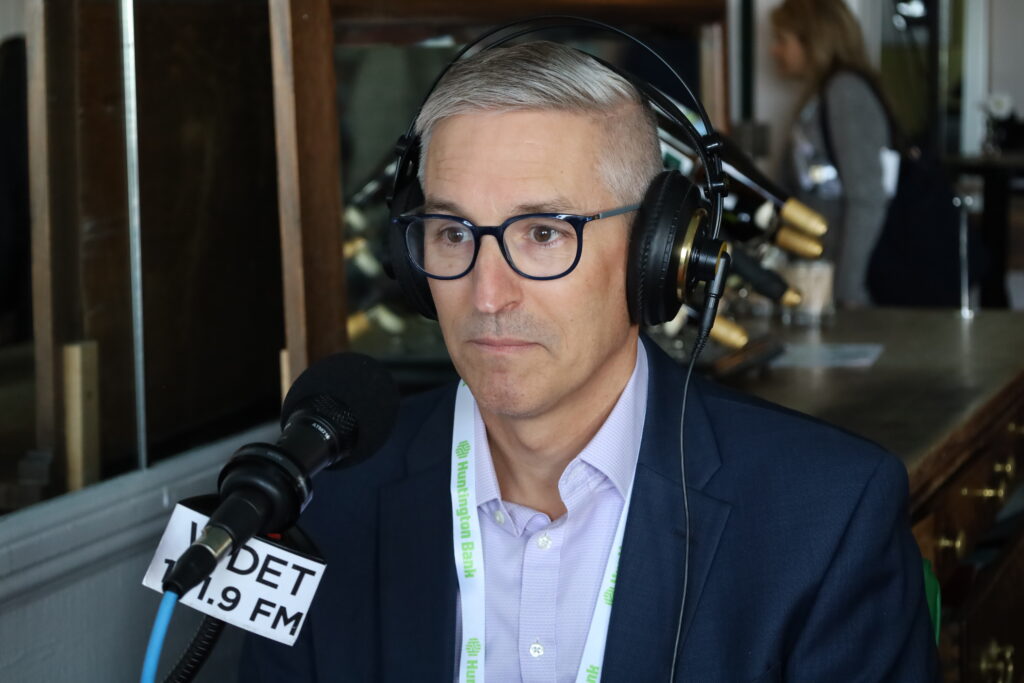 Joe Agostinelli sits in front of a WDET microphone with headphones on. He's wearing glasses and a light purple shirt with a black suit jacket.
