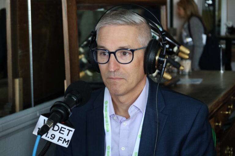 Joe Agostinelli sits in front of a WDET microphone with headphones on. He's wearing glasses and a light purple shirt with a black suit jacket.