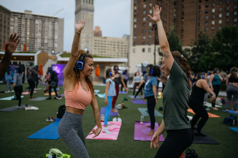Two women facing each other smile while stretching one arm up in a yoga pose. Behind them, you can see more people on yoga mats in the Campus Martius green.