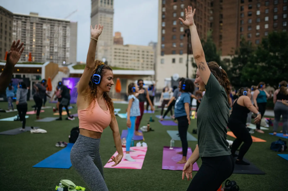 Two women facing each other smile while stretching one arm up in a yoga pose. Behind them, you can see more people on yoga mats in the Campus Martius green.