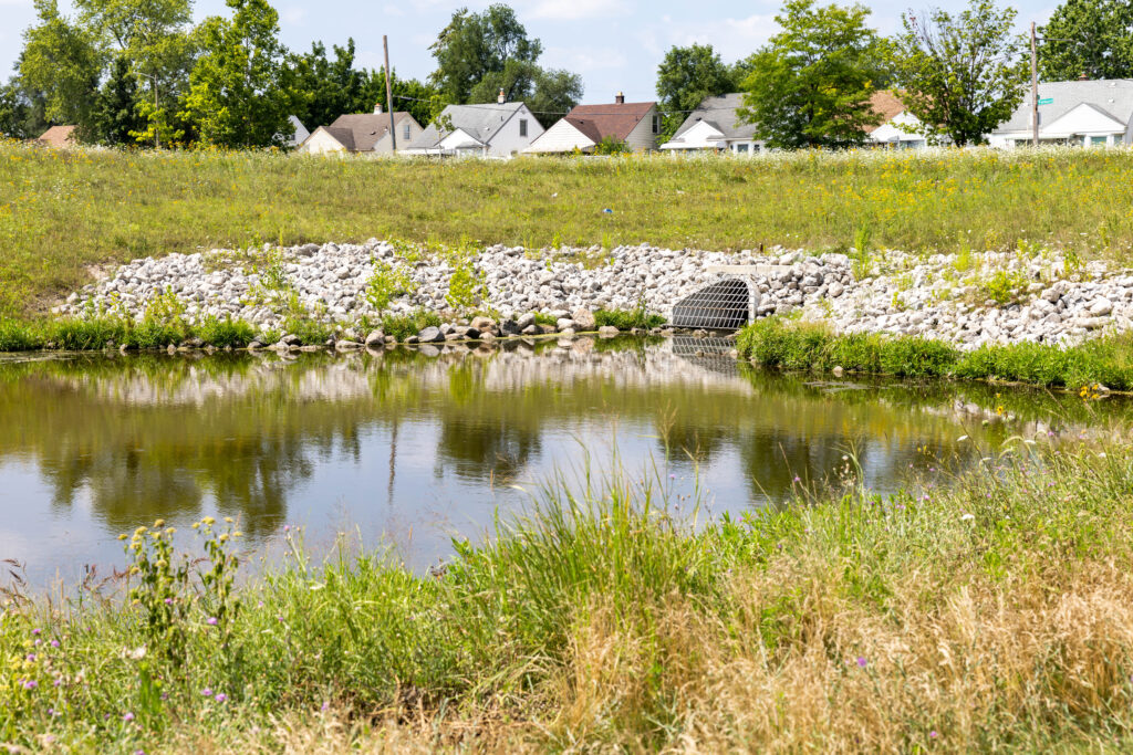A small body of clear water, filtered in part by a storm drain. Houses stand in the background.