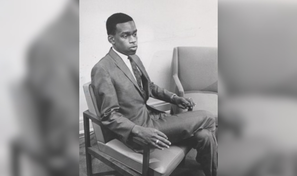 A black and white portrait of a young Jay Butler, sitting in a suit.
