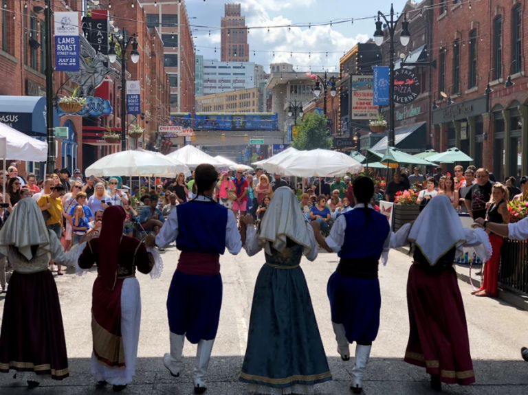 A line of traditional Greek dancers stands in front of an audience.