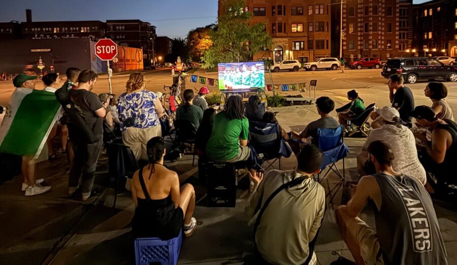 Around 15 people gather in front of a TV, sitting on lawn chairs and crates.