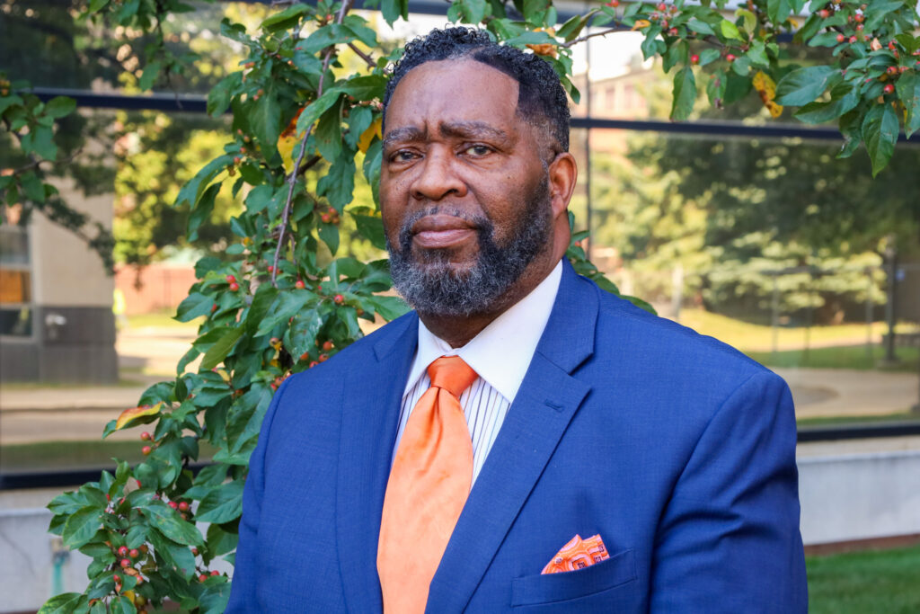 Bishop Williams stands in front of a leafy green tree wearing a blue suit and orange tie.