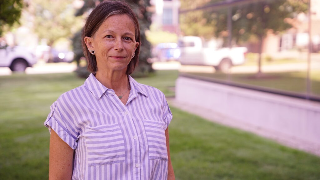 Pepper Catt wears a striped shirt, posed in front of trees and reflective glass outside of WDET.