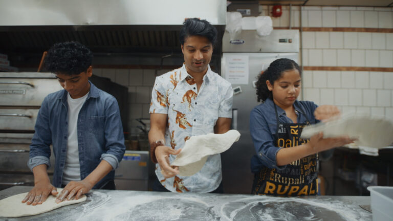 A father and his son and daughter stand in the kitchen of a pizza place working on dough.