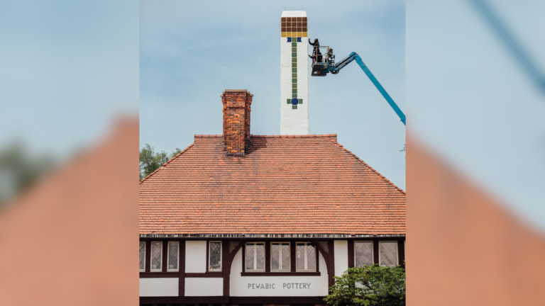 Two women paint tile-like patterns on Pewabic's chimney, held aloft by a blue construction lift.